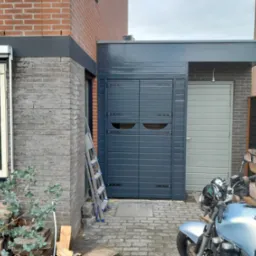 Nowoczesna wiata garażowa z dwuskrzydłowymi drzwiami w kolorze grafitowym, z widocznymi okuciami, przylegająca do budynku z cegły, z motocyklem zaparkowanym przed nią i drabiną opartą o ścianę.