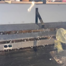 Kacper Dudra Stolarstwo-Montaż - Szare płytki ścienne z metalowymi listwami i krzyżykami dystansowymi w trakcie montażu nad czarnym blatem kuchennym ze zlewem. Widoczne otwory na gniazdka elektryczne i wsporniki półki.