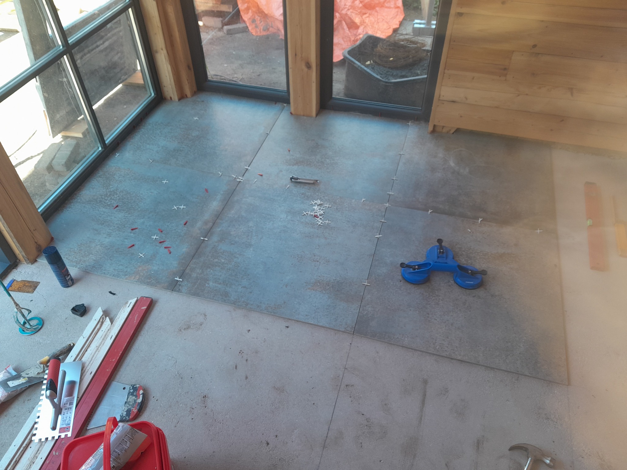 Układanie płytek podłogowych w nowym budynku, widoczne narzędzia glazurnicze, przyssawka do płytek, krzyżyki dystansowe i poziomica, prace w toku.