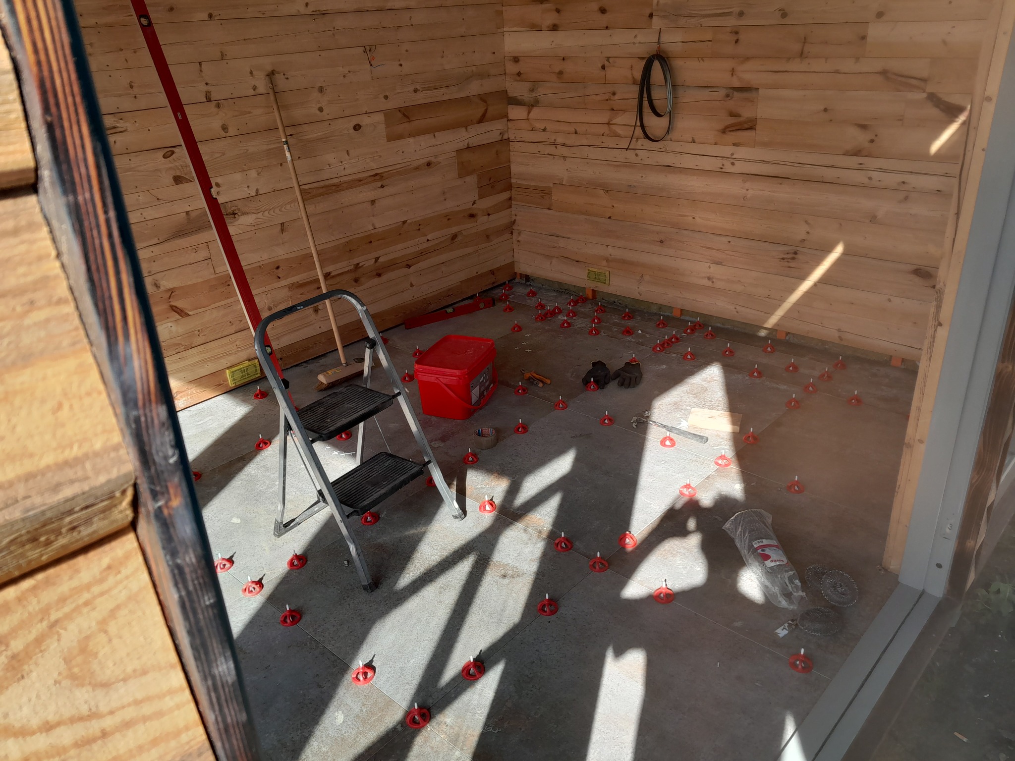 Wnętrze drewnianego domku z częściowo ułożoną szarą podłogą z płytek, wykorzystującą czerwone klipsy poziomujące. Na podłodze widoczne narzędzia, wiadro i drabina. Ściany wyłożone boazerią.