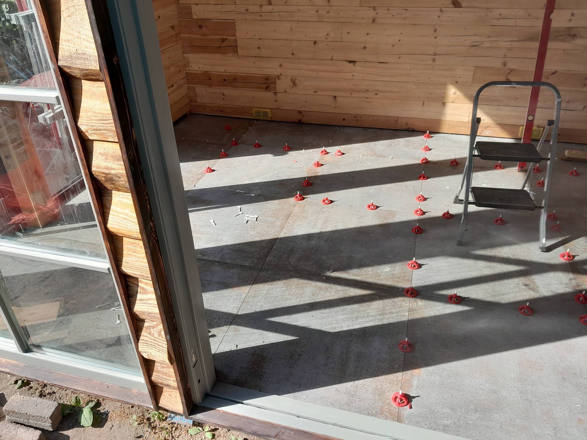 Proces układania płytek podłogowych w drewnianym domku, z użyciem poziomicy i czerwonych klipsów dystansowych, widoczne cienie i drabina.