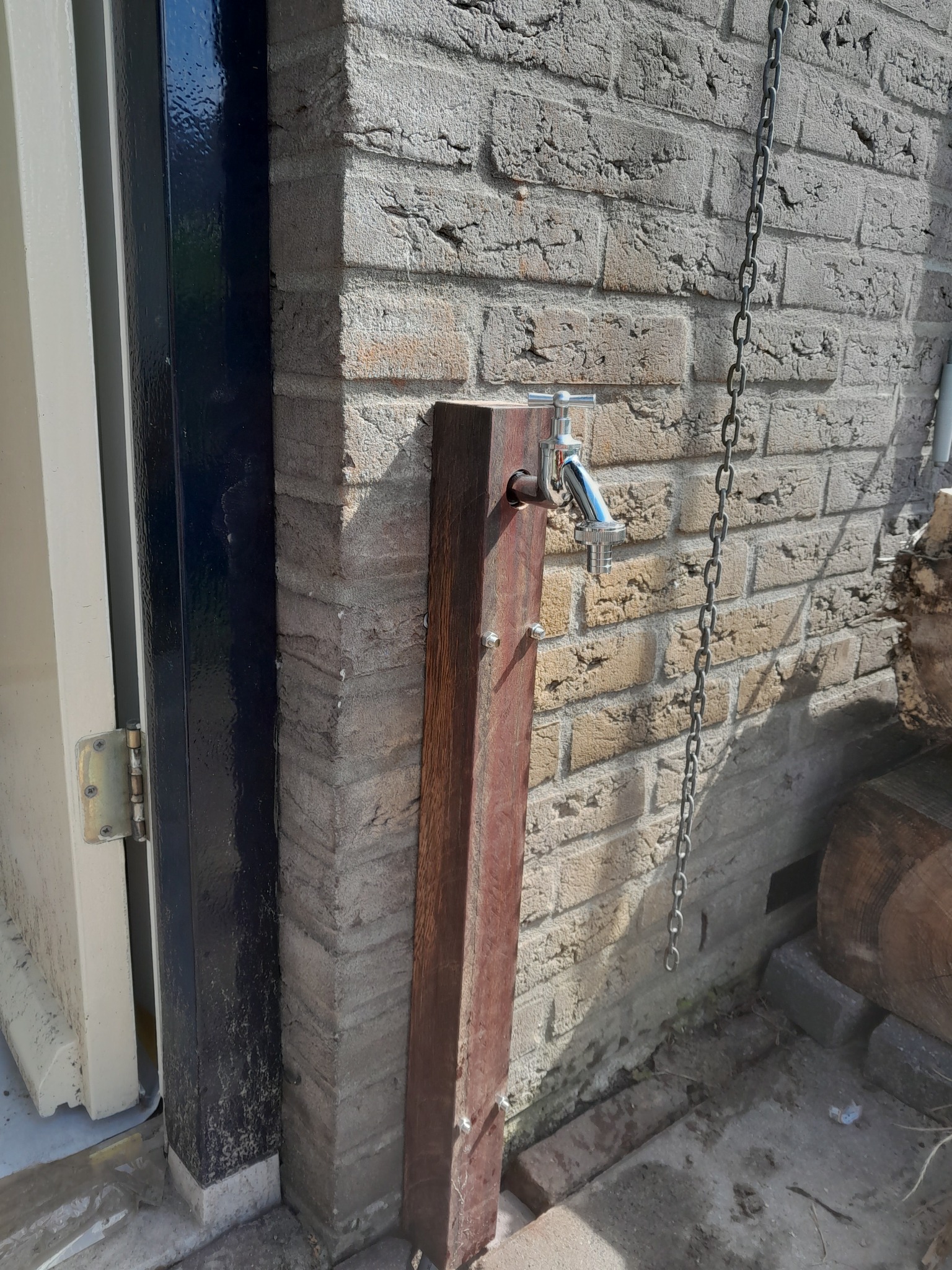 Zewnętrzny kran zamontowany na drewnianym słupku przy ceglanej ścianie, obok drzwi z czarną ramą i wiszącym łańcuchem.