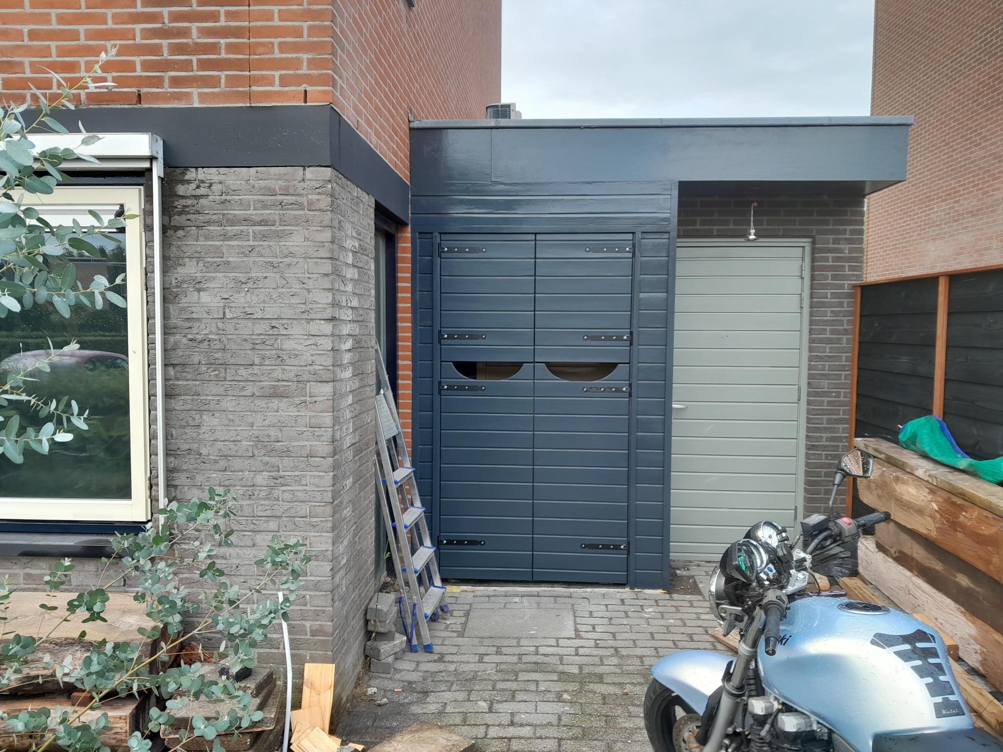 Nowoczesna wiata garażowa z dwuskrzydłowymi drzwiami w kolorze grafitowym, z widocznymi okuciami, przylegająca do budynku z cegły, z motocyklem zaparkowanym przed nią i drabiną opartą o ścianę.