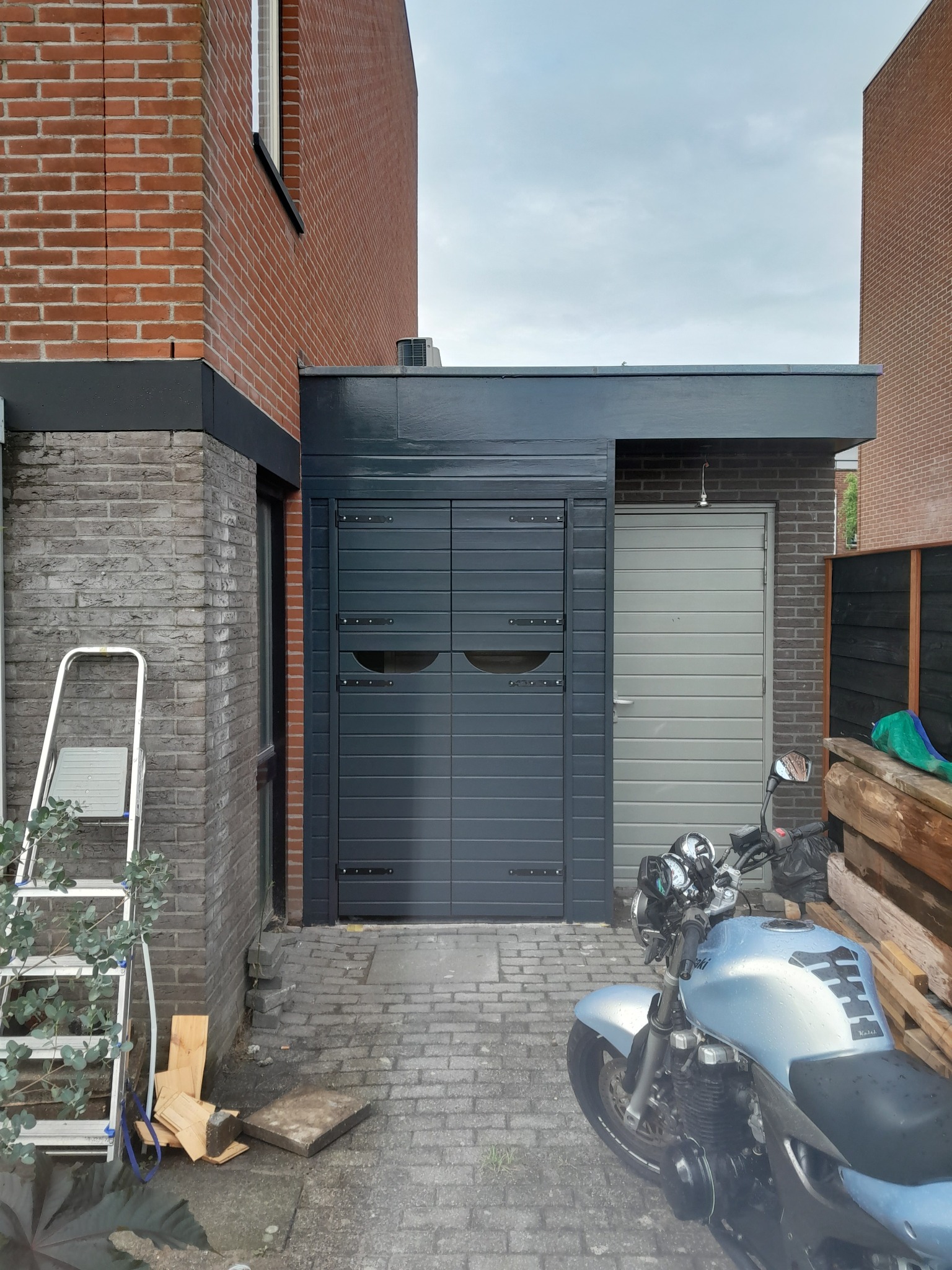 Szara, pozioma brama garażowa z dwoma skrzydłami, umieszczona w murowanej konstrukcji, obok szare drzwi wejściowe; na pierwszym planie częściowo widoczny motocykl, obok drabina i materiały budowlane.