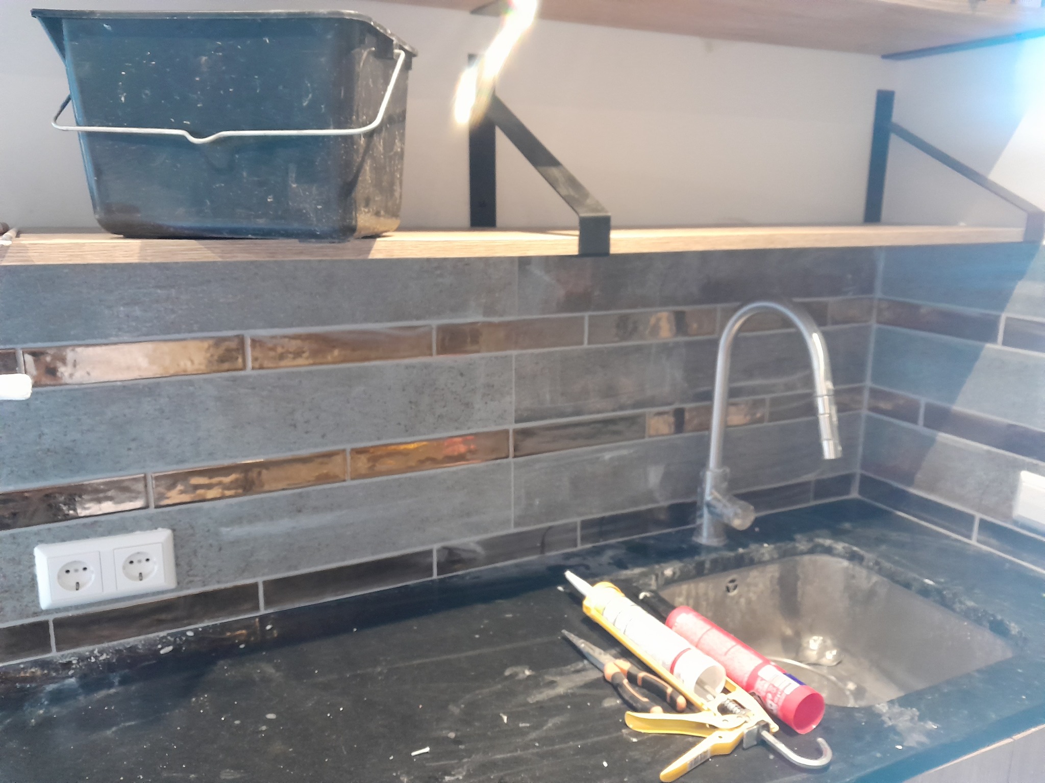 Ułożona glazura w kuchni z fugami, widoczny zlew, bateria, gniazdko elektryczne, wiadro i narzędzia do silikonowania.