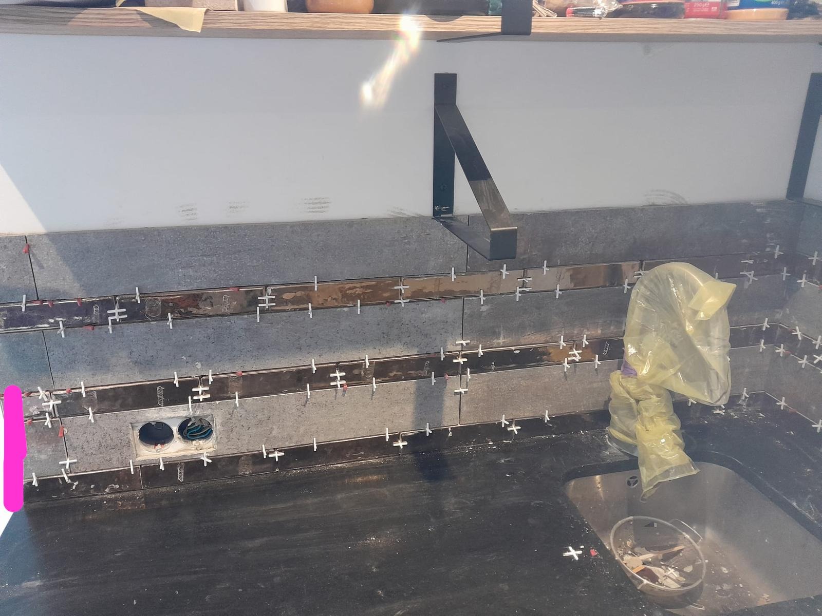 Szare płytki ścienne z metalowymi listwami i krzyżykami dystansowymi w trakcie montażu nad czarnym blatem kuchennym ze zlewem. Widoczne otwory na gniazdka elektryczne i wsporniki półki.