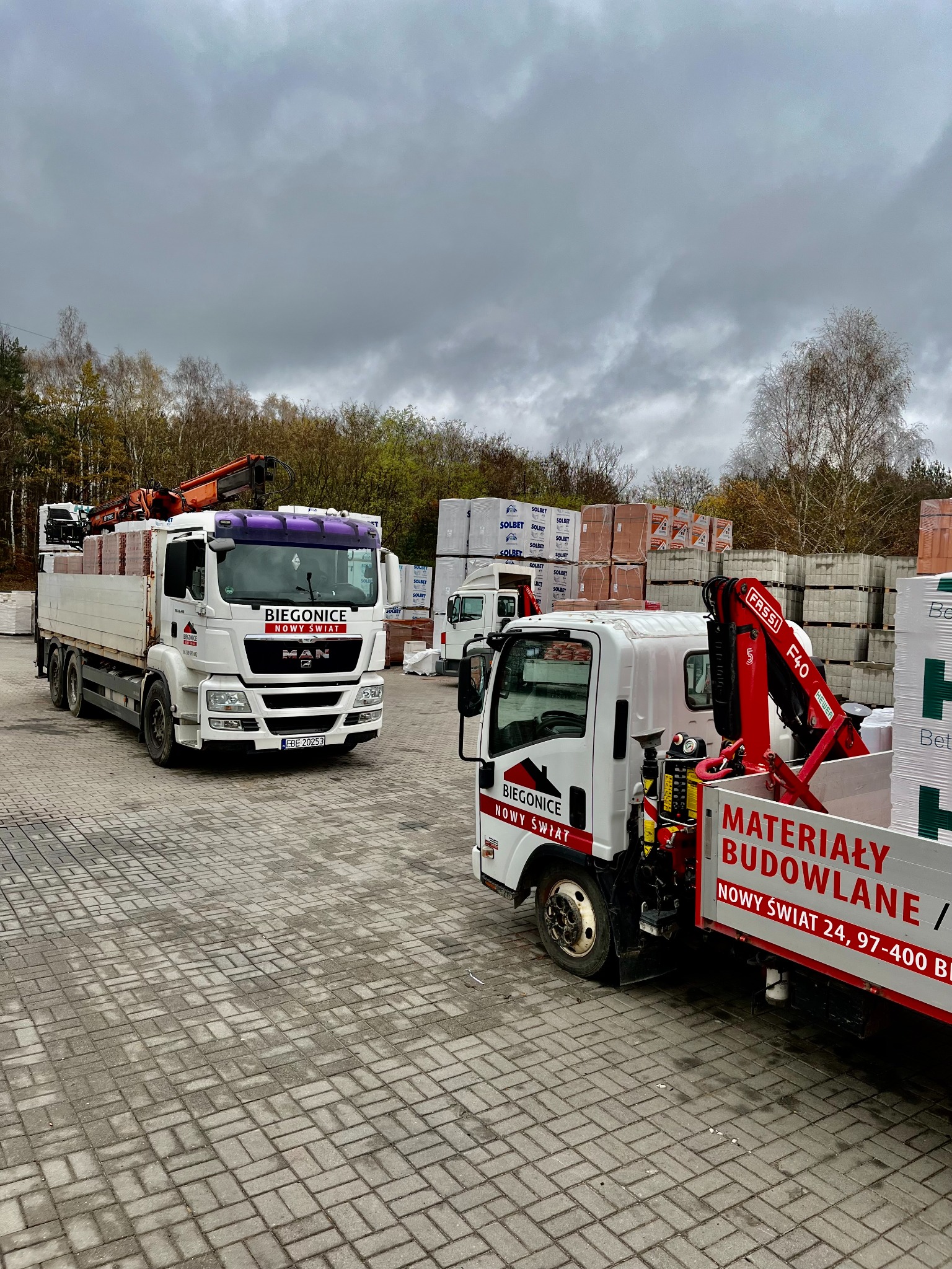 Dwa samochody dostawcze z dźwigami, załadowane materiałami budowlanymi, stoją na placu wyłożonym kostką brukową; na samochodach widoczne logo firmy 'Biegonice Nowy Świat'.