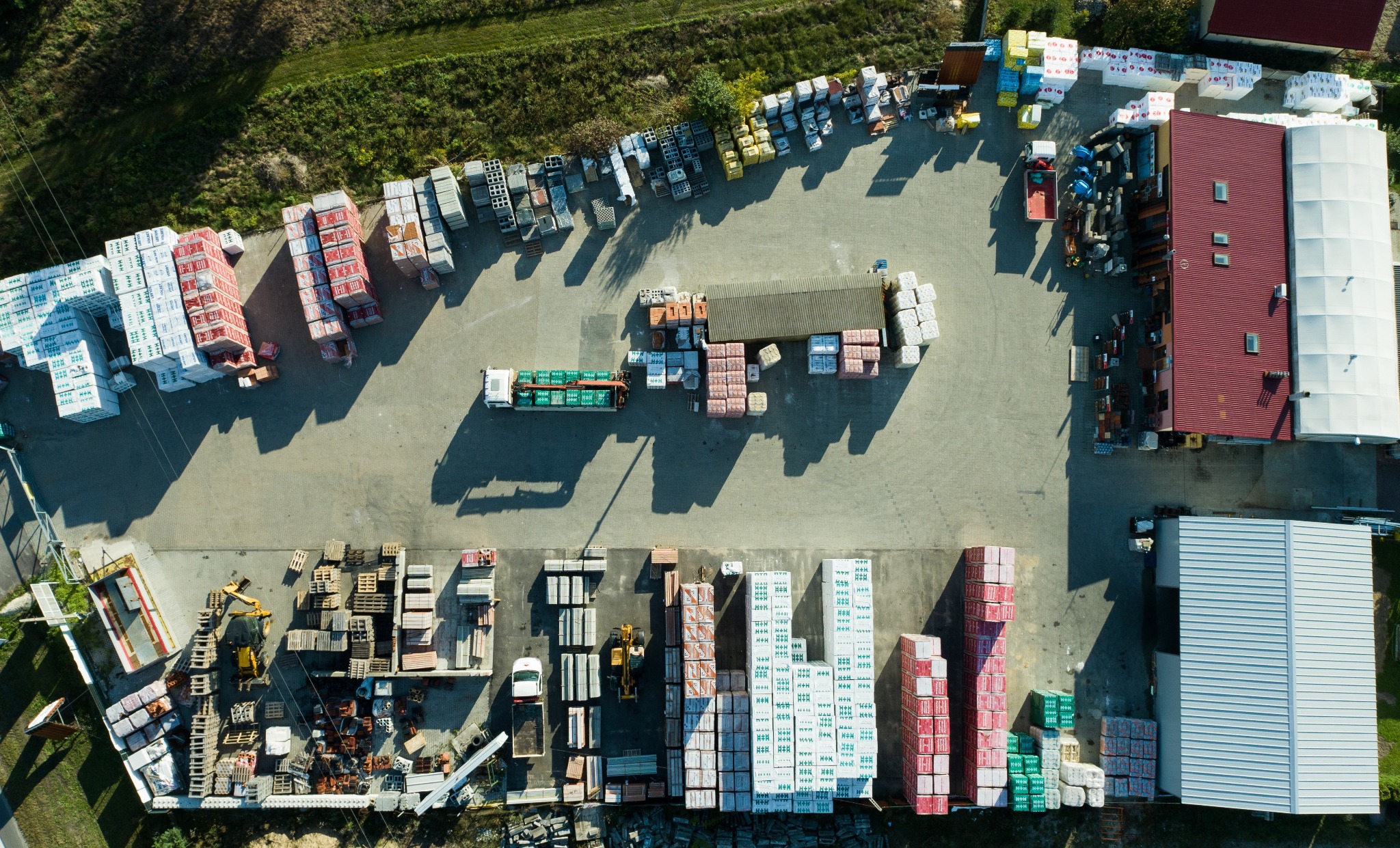 Widok z lotu ptaka na plac budowlany z zapasem materiałów budowlanych, w tym palety z bloczkami, transport ciężarowy oraz budynek magazynowy z czerwonym dachem.
