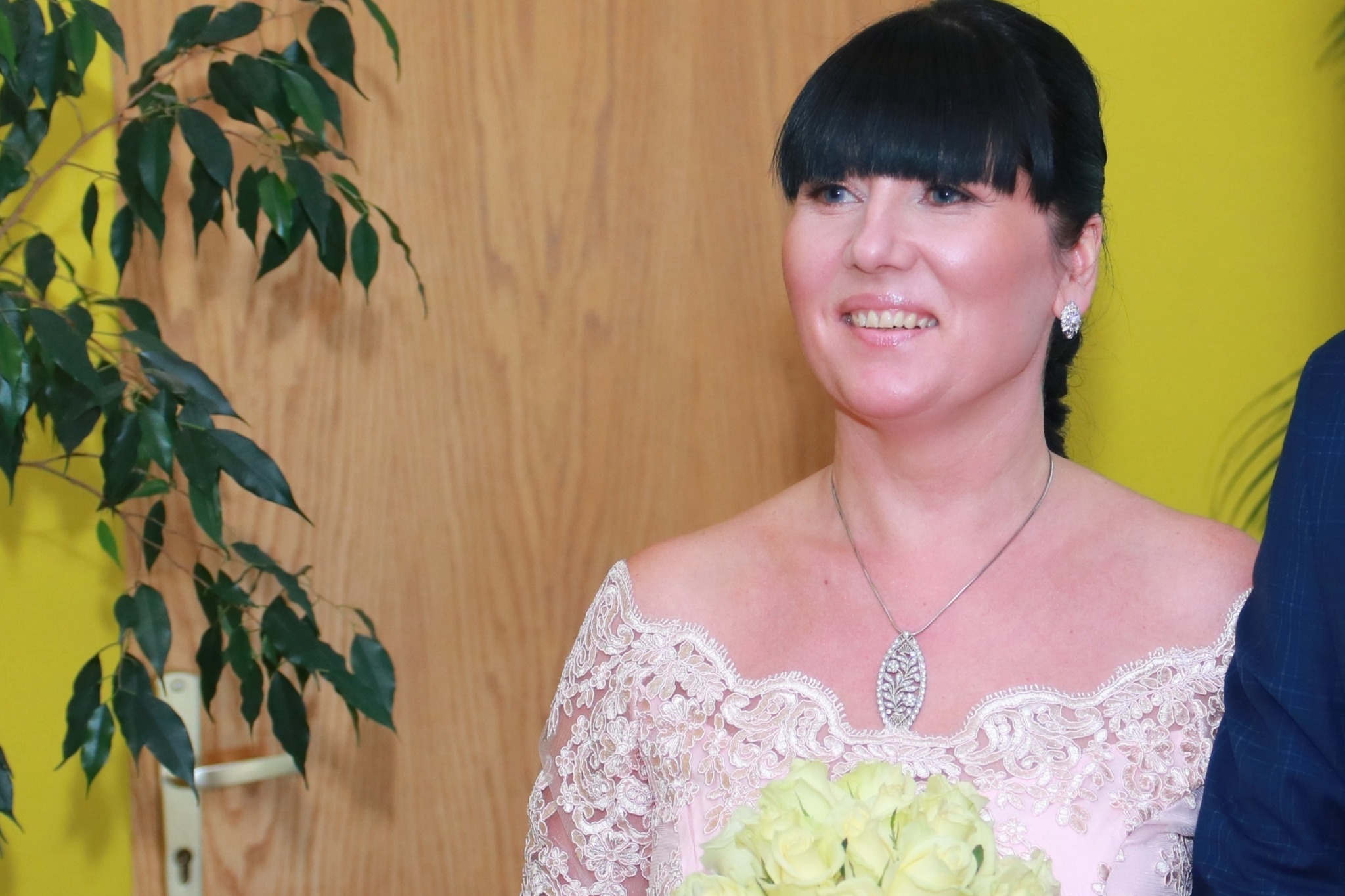 Portret uśmiechniętej kobiety w koronkowej sukience z bukietem bladożółtych róż, na tle drewnianej ściany i zielonej rośliny doniczkowej.