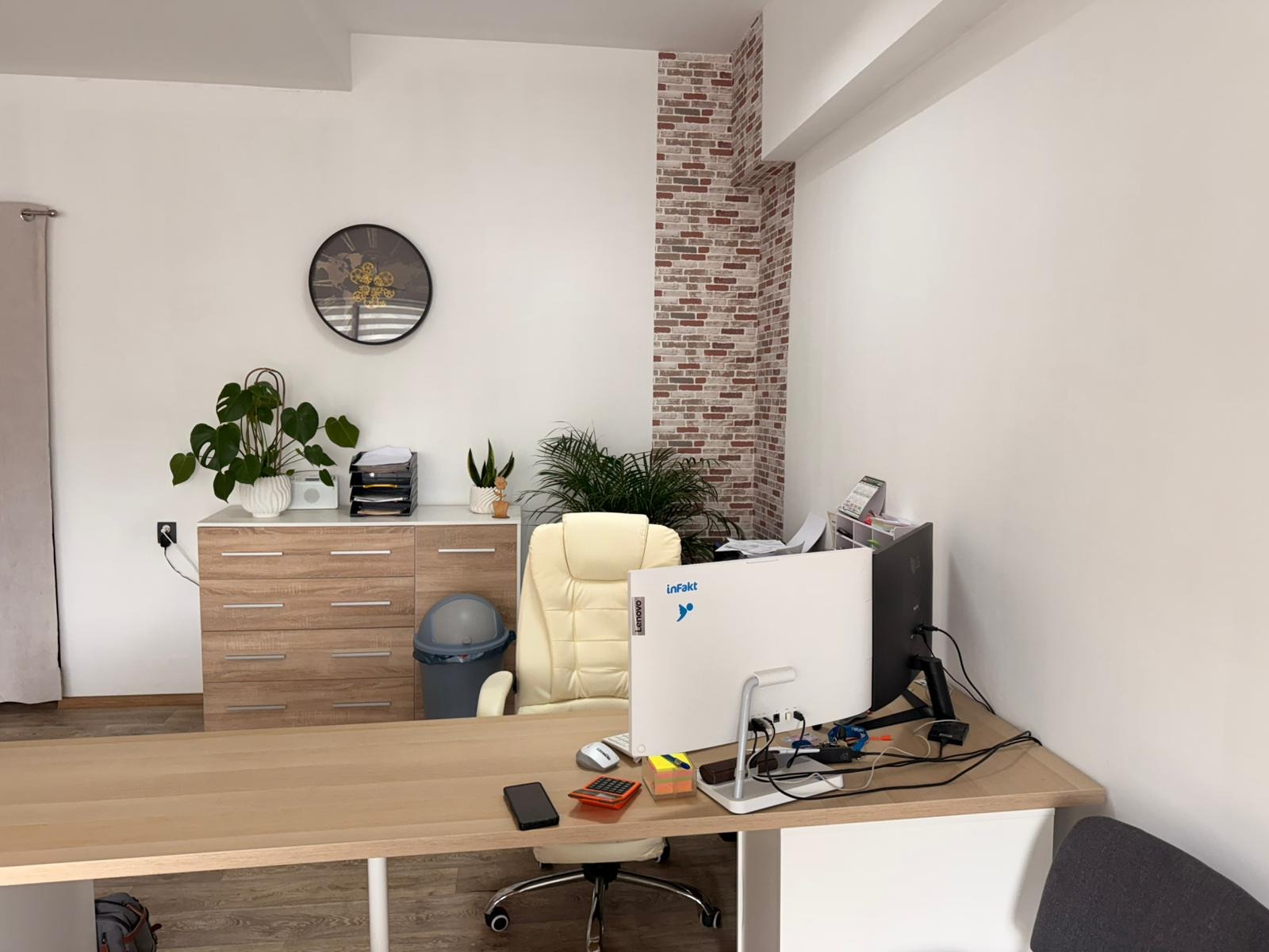Jasne biuro z biurkiem, komputerem i roślinami. Na ścianie zegar i dekoracyjna cegła. Widok na uporządkowane stanowisko pracy.