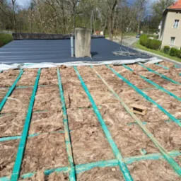 Ocieplenie poddasza wełną mineralną, widoczne turkusowe belki konstrukcyjne, fragment pokrytego blachą dachu w tle, budynek mieszkalny i drzewa w tle.