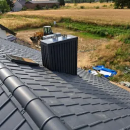 Nowy, grafitowy dach z dachówki modułowej, widoczne okno dachowe, komin w obudowie z blachy trapezowej, w tle krajobraz wiejski z budynkami i polem, na podwórzu koparka.