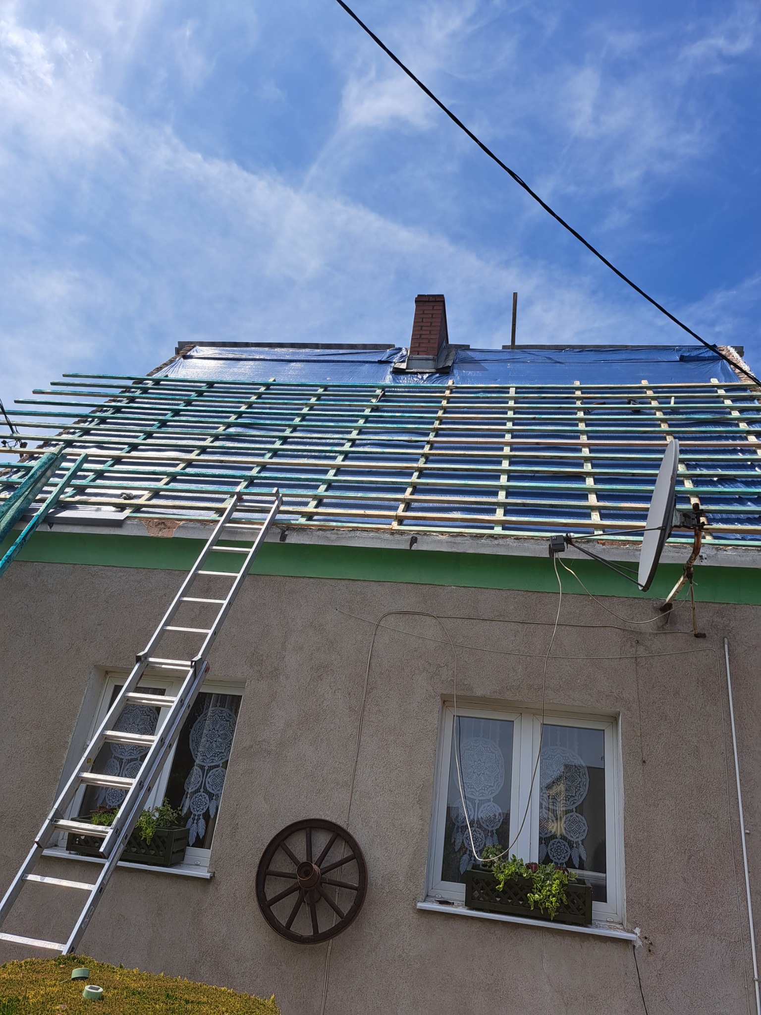 Dom z niebieską folią na dachu, na którym ułożone są drewniane łaty, drabina oparta o ścianę, widoczny komin i antena satelitarna.