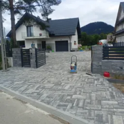 Ułożona szara kostka brukowa na podjeździe do nowoczesnego domu z widokiem na góry, ubijarka wibracyjna i akcesoria brukarskie obok.