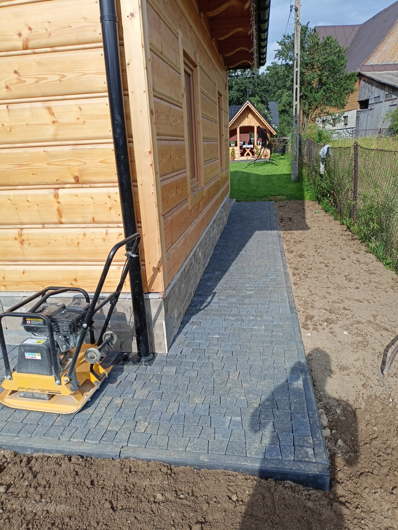 Świeżo ułożona, szara kostka brukowa obok drewnianego domu z widoczną zagęszczarką płytową, trawnikiem i altaną w tle.
