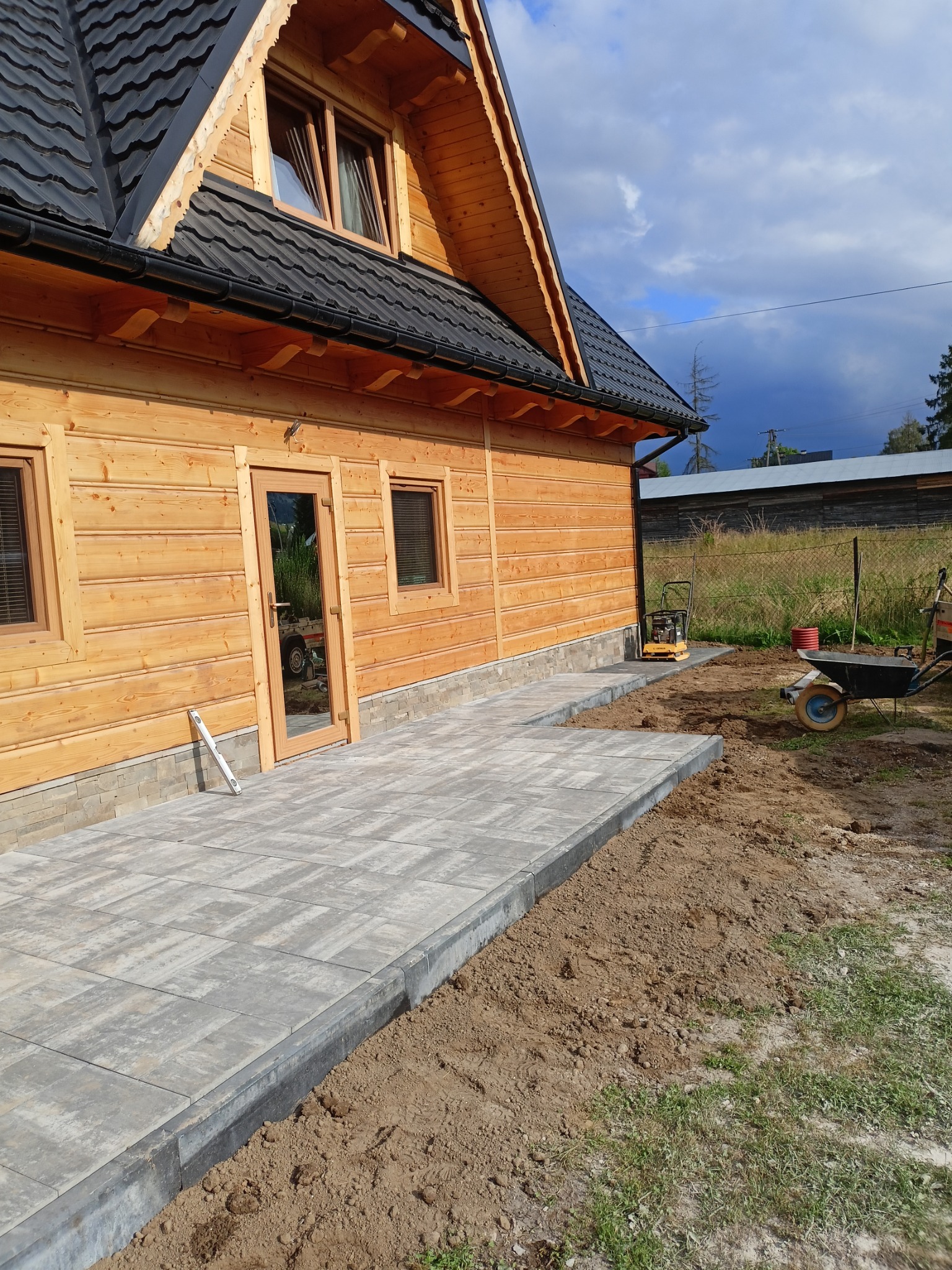 Nowo ułożony chodnik z szarych płyt betonowych prowadzący do drewnianego domu, z widoczną zagęszczarką i taczką w tle, oraz poziomicą opartą o ścianę domu.