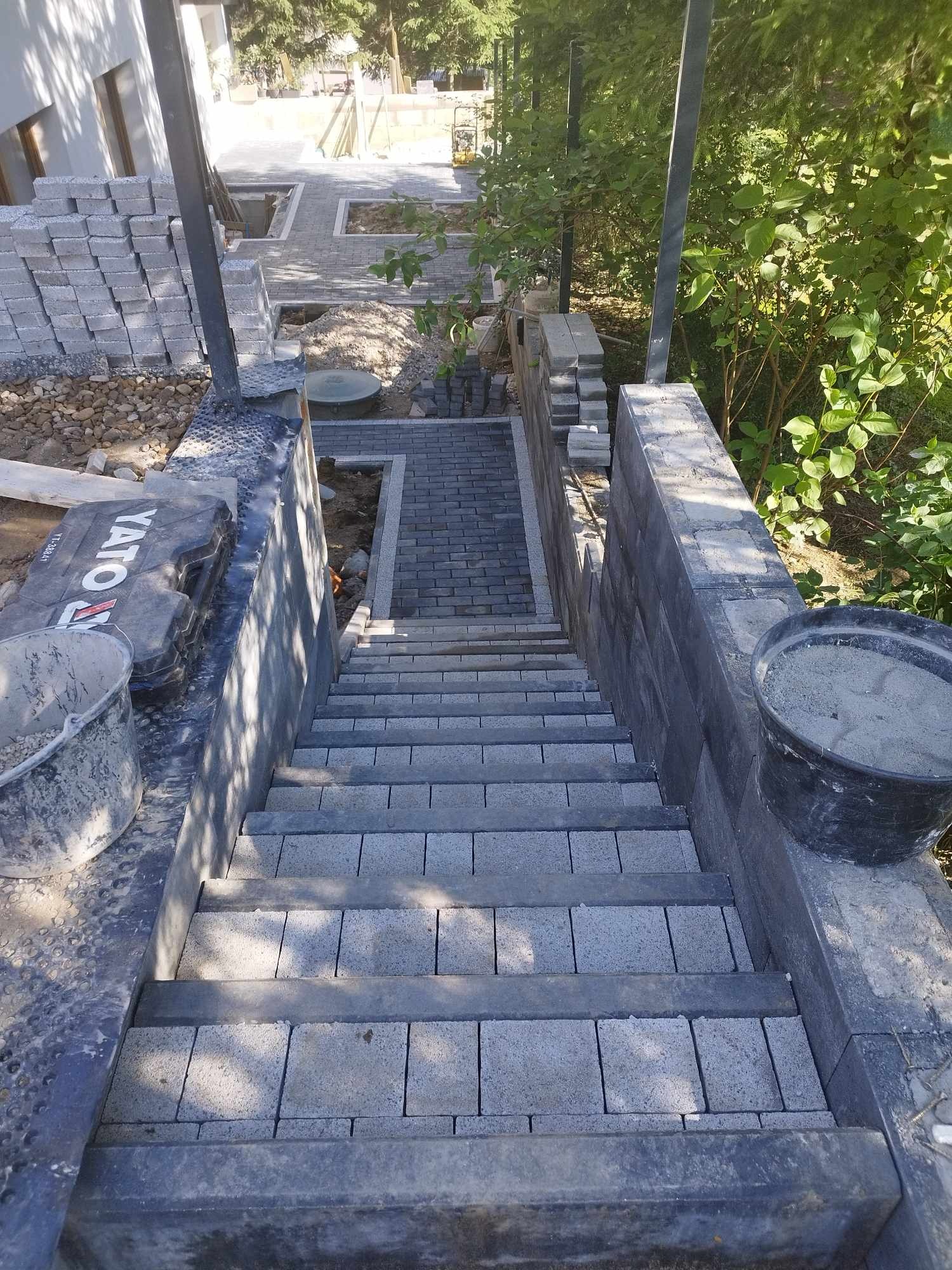 Schody z kostki brukowej w trakcie budowy, widok z góry, z widocznymi narzędziami i materiałami budowlanymi, w tle ułożona ścieżka.