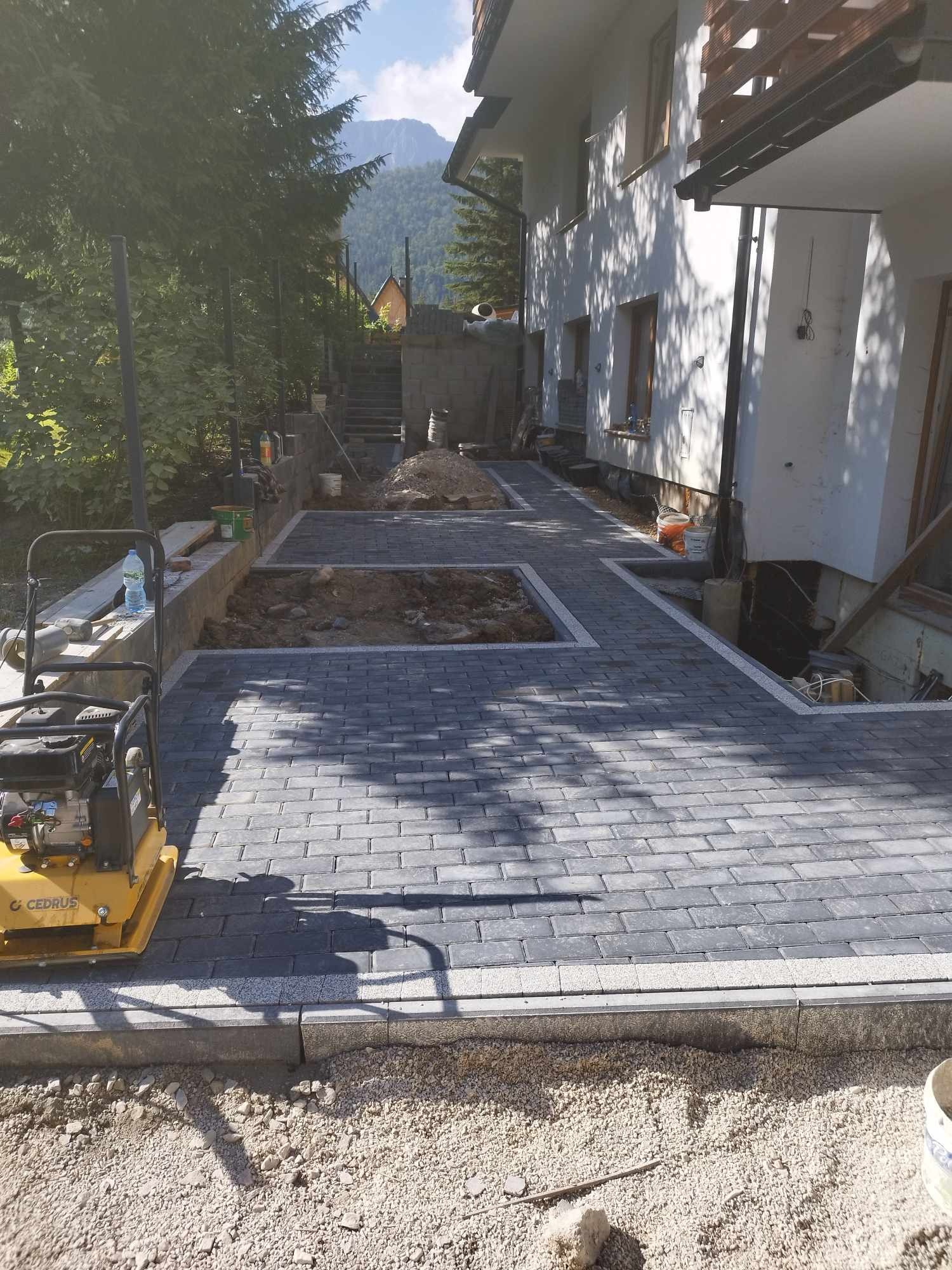 Świeżo ułożony chodnik z szarej kostki brukowej przy białym budynku, z żółtą zagęszczarką w rogu i widokiem na góry w tle.