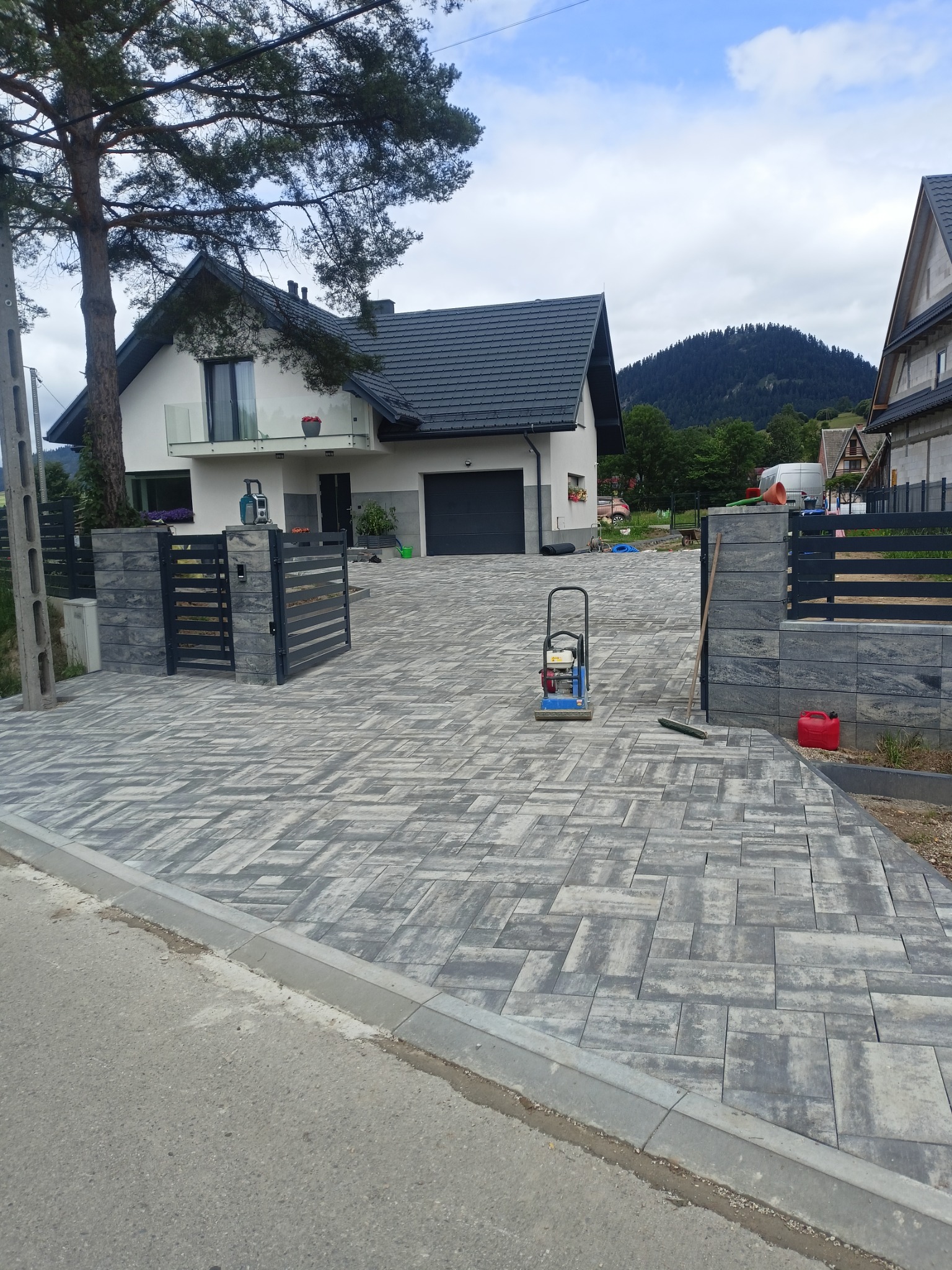 Ułożona szara kostka brukowa na podjeździe do nowoczesnego domu z widokiem na góry, ubijarka wibracyjna i akcesoria brukarskie obok.