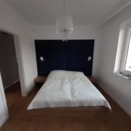 Nowoczesna sypialnia z drewnianą podłogą, łóżkiem z tapicerowanym, granatowym zagłówkiem i wiszącą lampą z kryształkami. Po bokach łóżka znajdują się drewniane szafki nocne z małymi lampkami. Widok...