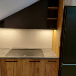 Fragment kuchni z zabudową pod skosem, drewniane szafki dolne i czarne górne, płyta indukcyjna na blacie, oświetlenie podszafkowe, lodówka w zabudowie po prawej stronie.