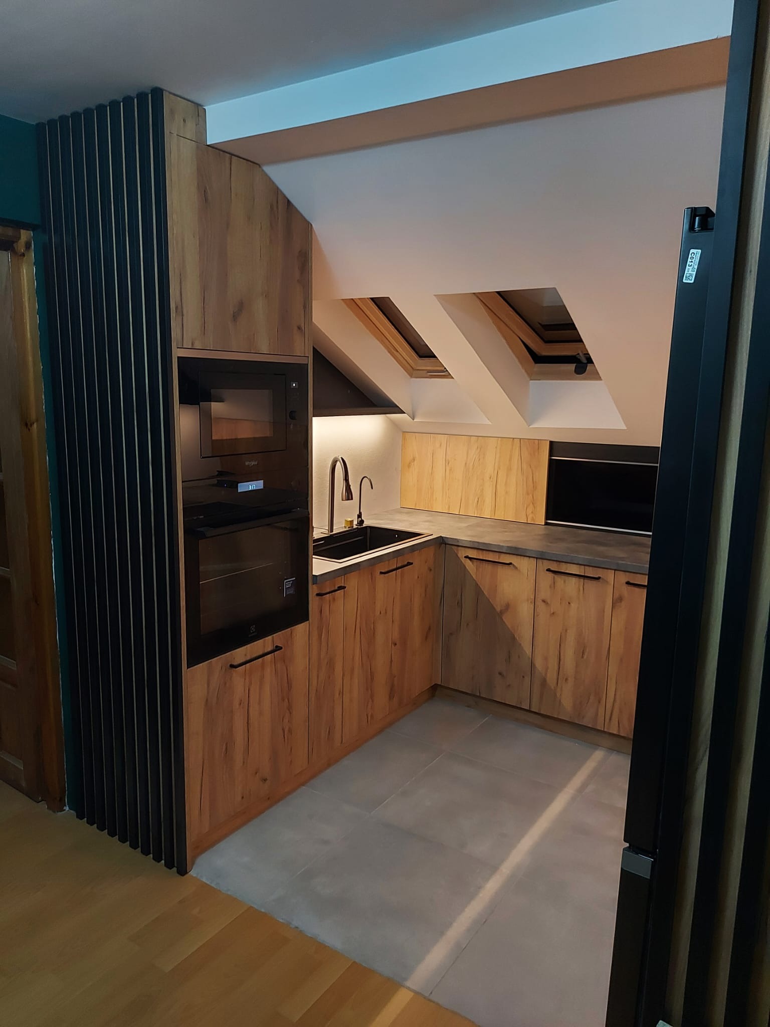 Kuchnia w stylu rustykalnym, zabudowa z drewnopodobnych frontów, czarny zlew i bateria, wbudowane sprzęty AGD, okna dachowe w skosach sufitu.