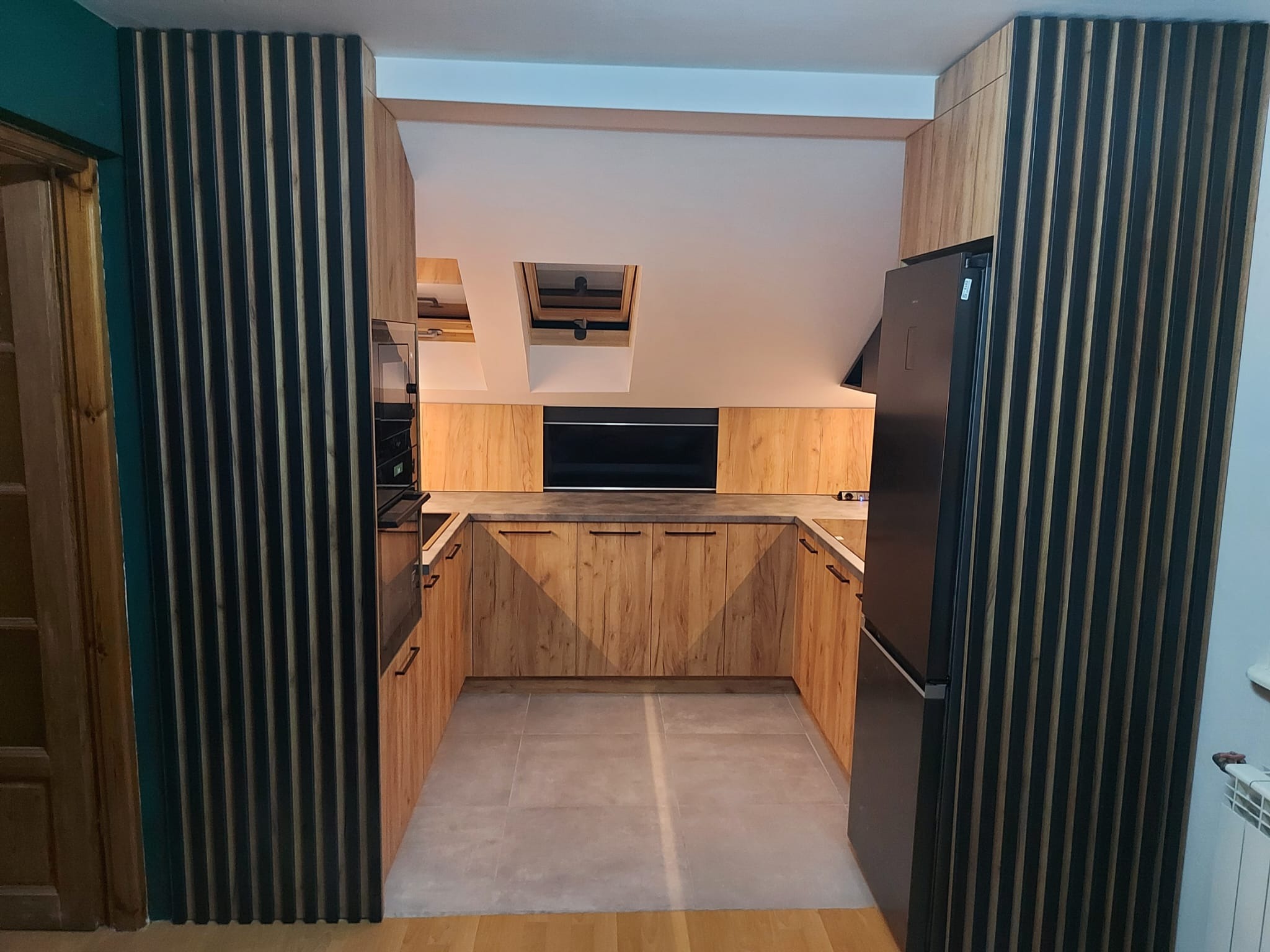 Nowoczesna kuchnia w kształcie litery U z drewnianymi frontami i szarym blatem, oświetlona światłem dziennym z okna dachowego, z czarnymi lamelami dekoracyjnymi po bokach.