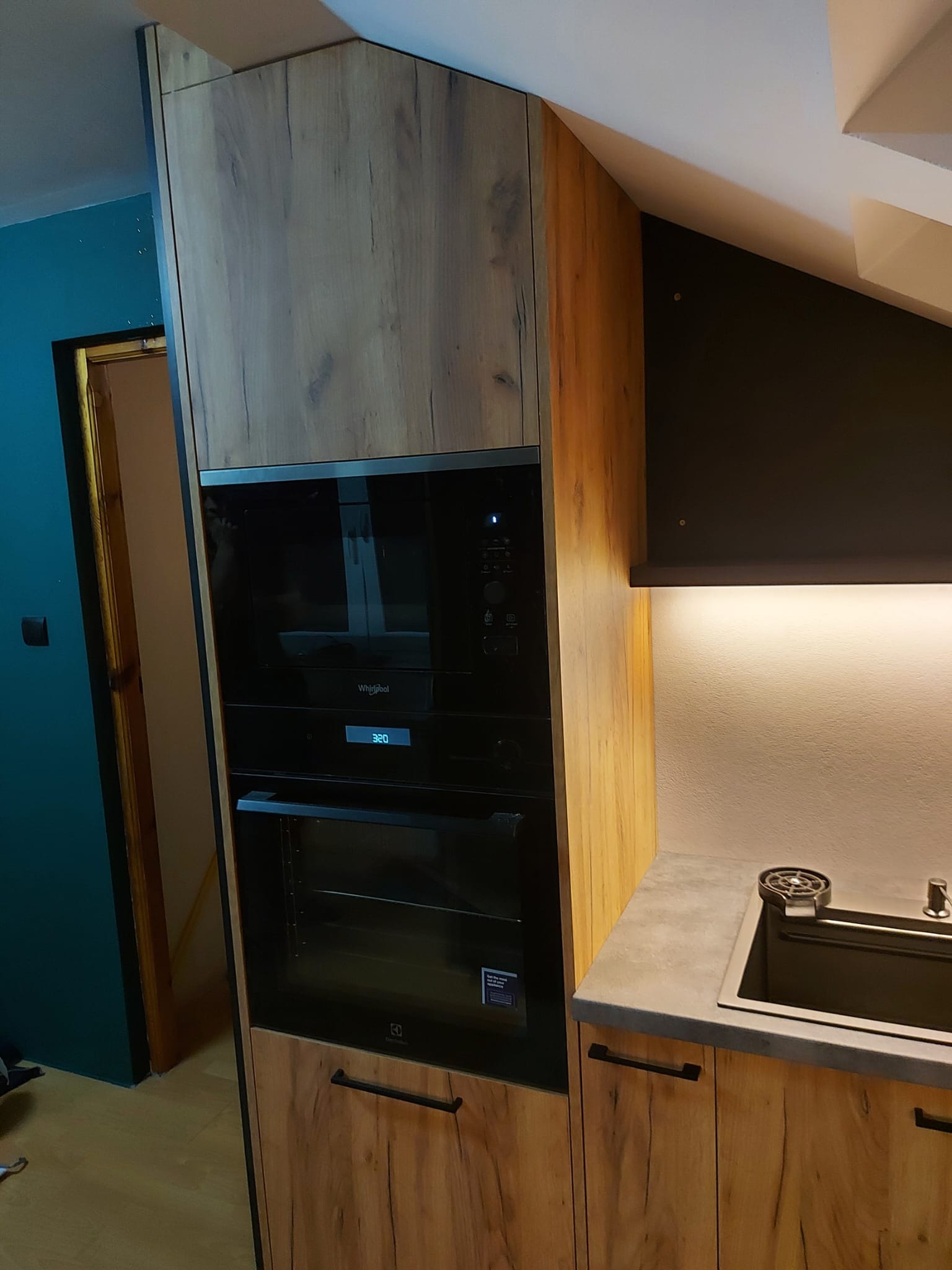 Nowoczesna zabudowa kuchenna z fornirem imitującym drewno, zintegrowana z piekarnikiem i mikrofalówką, podświetlony blat roboczy z szarym zlewem.