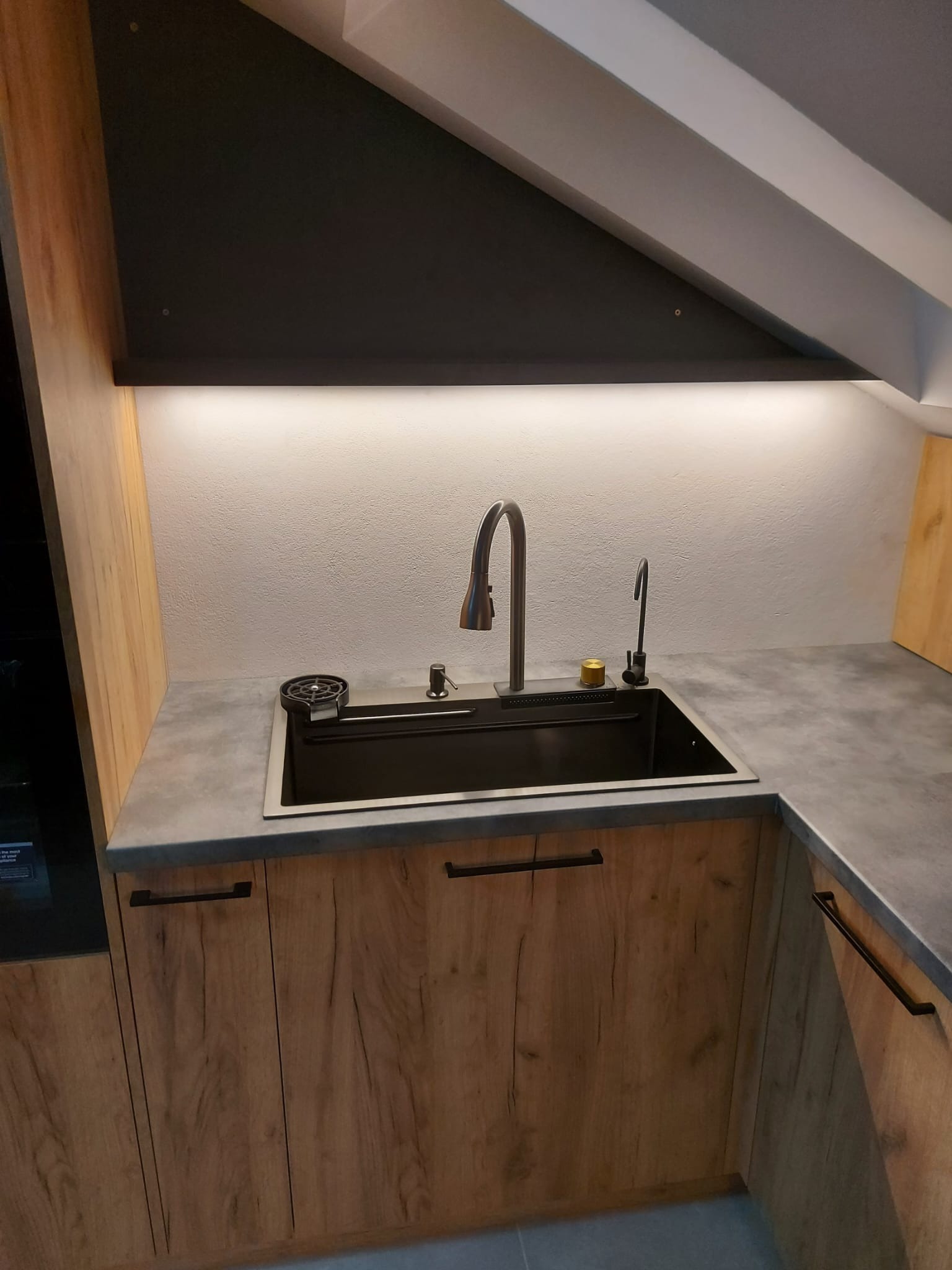 Nowoczesna kuchnia w skosie poddasza: czarny zlew, bateria i dozownik z szarym blatem, drewniane szafki z czarnymi uchwytami, oświetlenie LED podszafkowe.