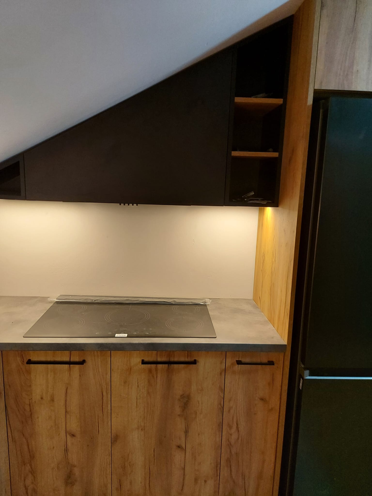 Fragment kuchni z zabudową pod skosem, drewniane szafki dolne i czarne górne, płyta indukcyjna na blacie, oświetlenie podszafkowe, lodówka w zabudowie po prawej stronie.