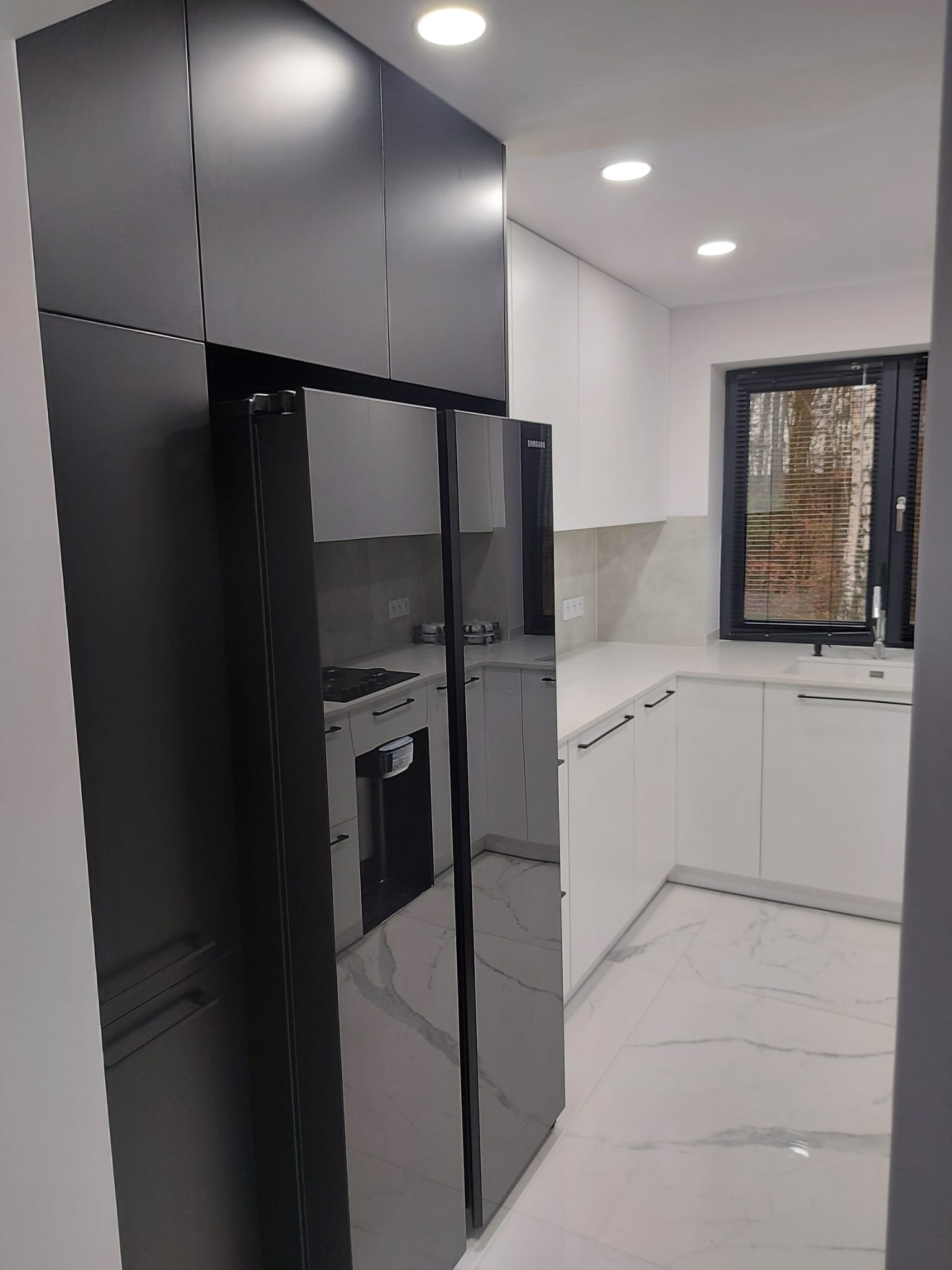 Nowoczesna kuchnia: czarne szafki, białe blaty, marmurowa podłoga. Lodówka Samsung z lustrzanym frontem odbija wnętrze. Okno z czarną ramą.