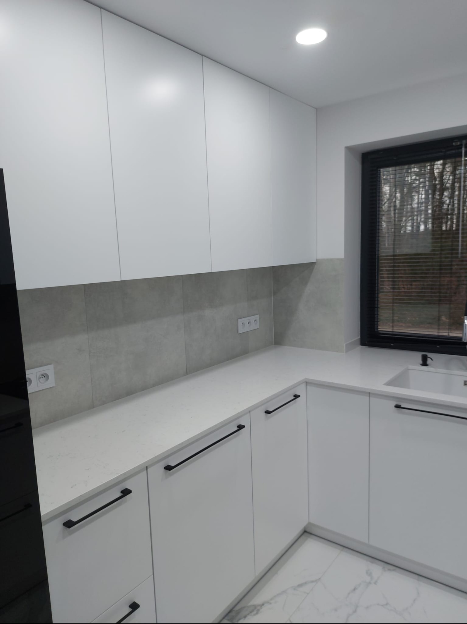 Nowoczesna, minimalistyczna kuchnia w bieli z czarnymi, podłużnymi uchwytami. Jasne blaty i marmurowe płytki podłogowe. Okno z czarną ramą.