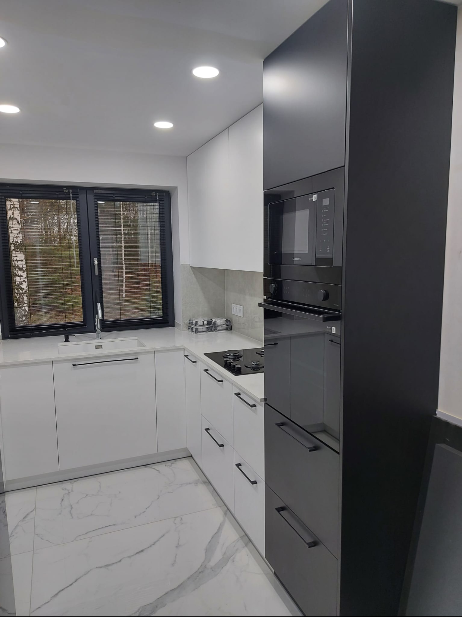 Nowoczesna kuchnia z białymi szafkami i czarnym wysokim modułem z piekarnikiem i mikrofalówką. Marmurowe płytki podłogowe i okno z żaluzjami.