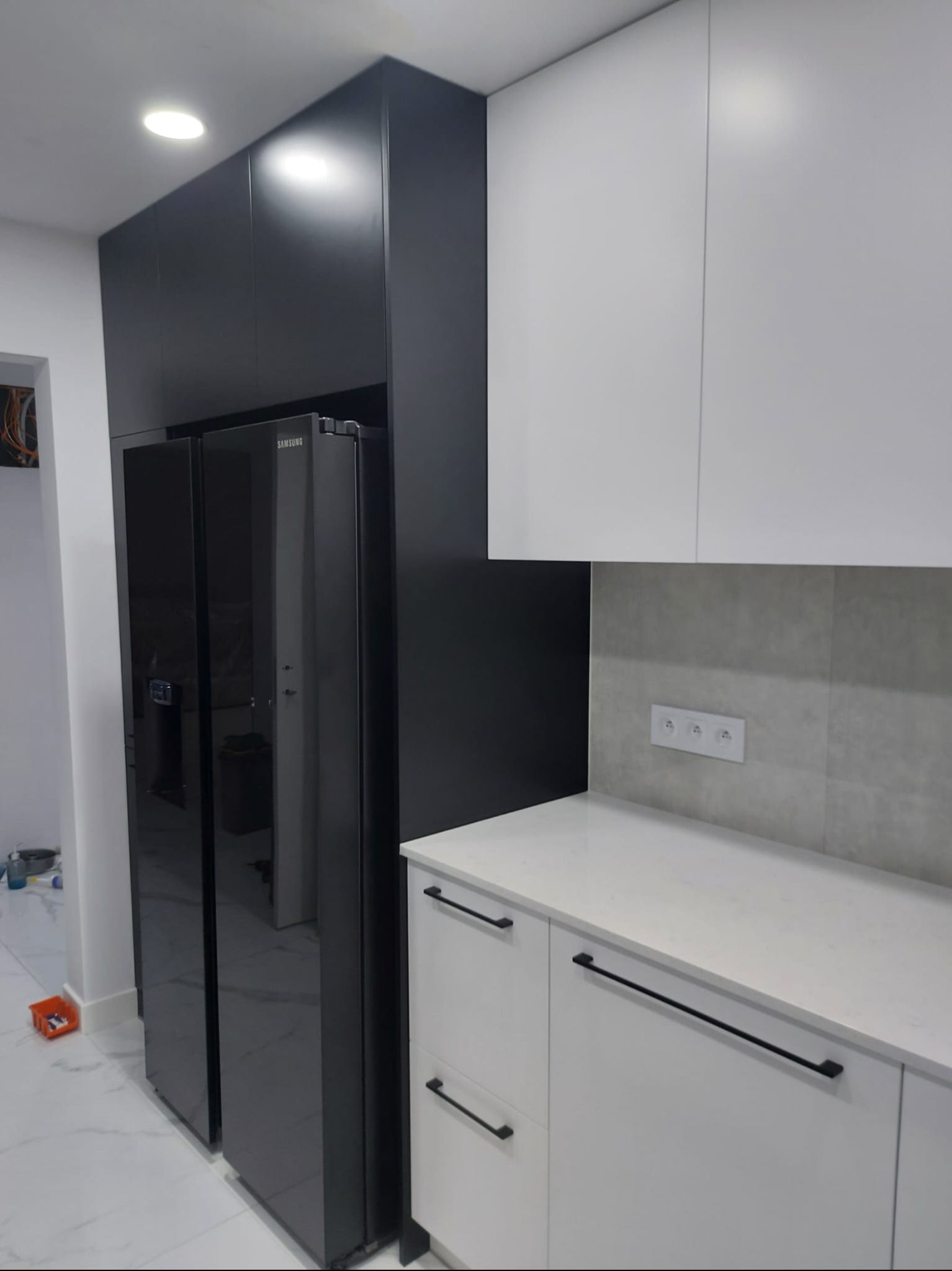 Nowoczesna kuchnia z czarną zabudową lodówki Samsung i białymi szafkami z czarnymi uchwytami. Minimalistyczny design, szara ściana nad blatem.