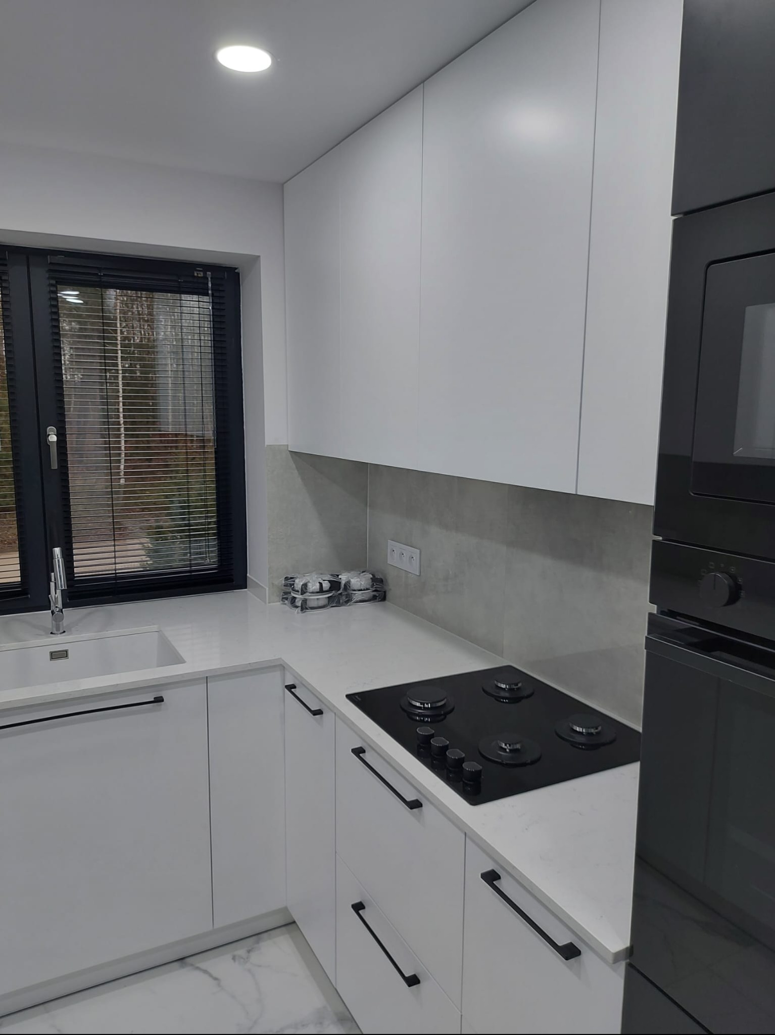Nowoczesna kuchnia z białymi, matowymi frontami, czarnymi uchwytami i płytą grzewczą. Marmurowa podłoga i blat. Okno z żaluzjami w czarnej ramie.