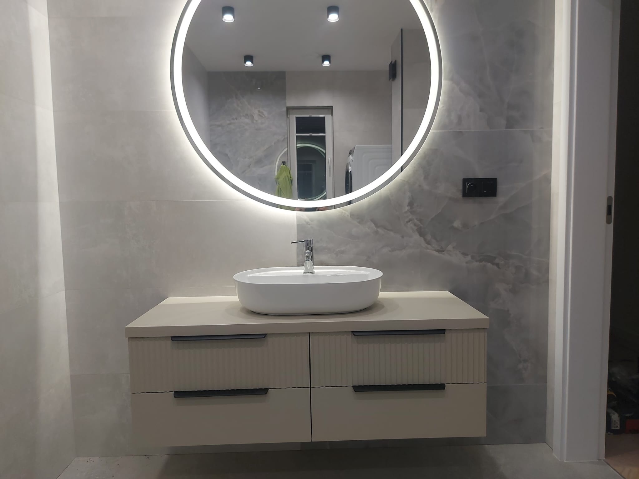 Nowoczesna łazienka z podświetlanym lustrem i szafką pod umywalką w odcieniach beżu. Na ścianie płytki imitujące marmur. Widoczne oświetlenie sufitowe.