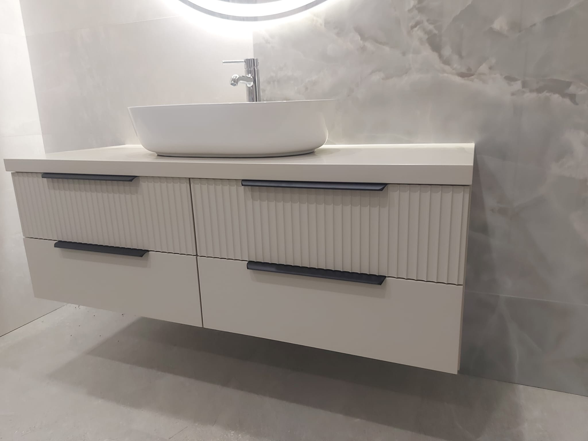 Beżowa szafka łazienkowa pod umywalkę nablatową z frezowanymi frontami i czarnymi uchwytami, na tle marmurowej ściany i okrągłego lustra z podświetleniem.