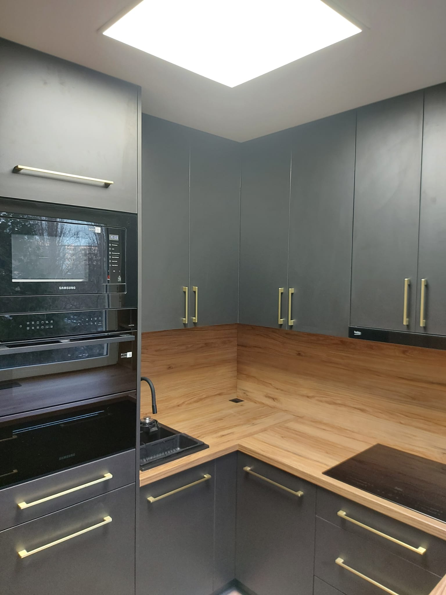 Nowoczesna kuchnia w odcieniach szarości z drewnianym blatem i złotymi uchwytami. Widoczny zlew, płyta indukcyjna i zabudowany sprzęt AGD.