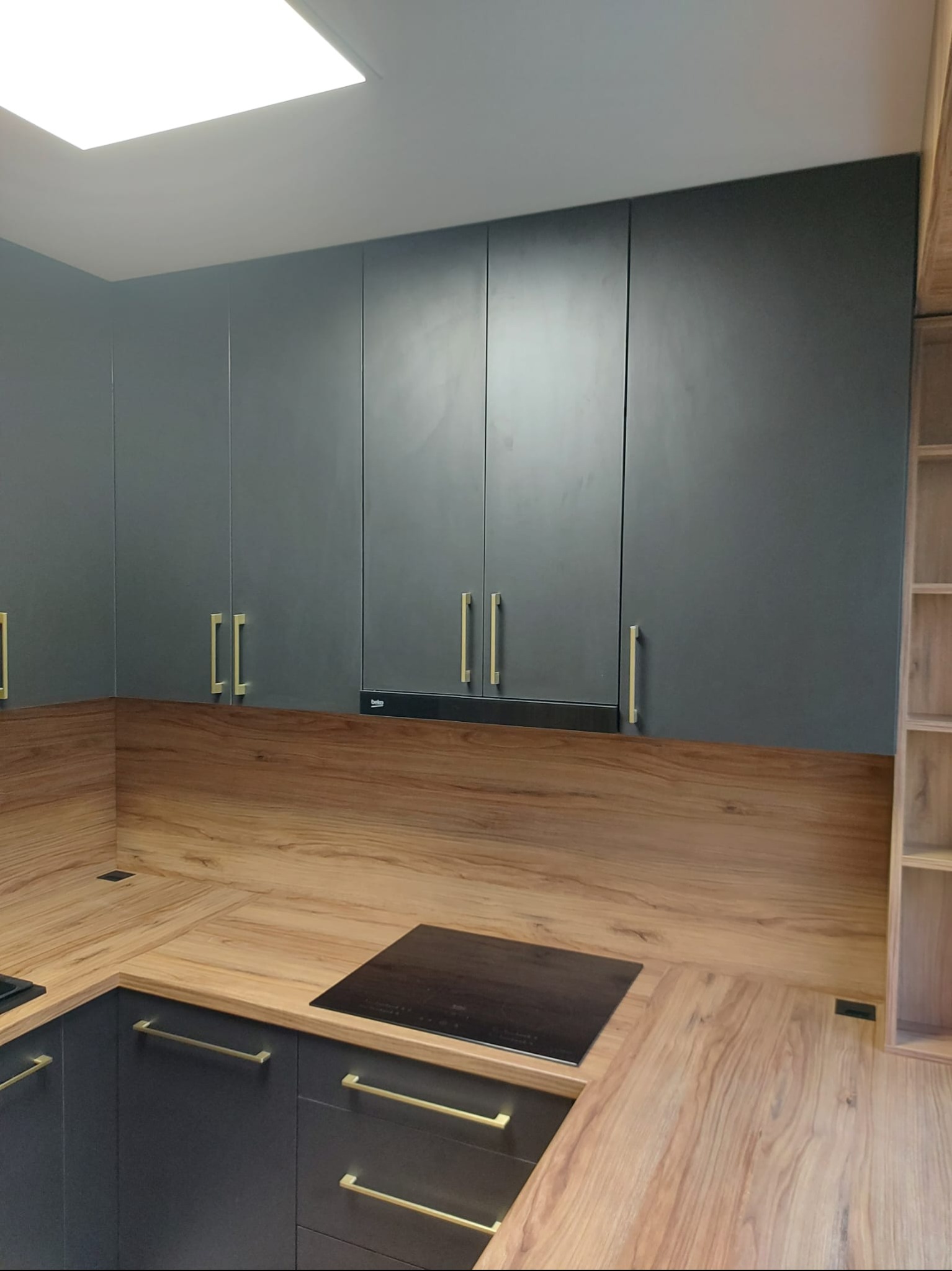 Nowoczesna kuchnia z szarymi szafkami i drewnianym blatem. Złote uchwyty dodają elegancji. Widoczna płyta indukcyjna i oświetlenie sufitowe.