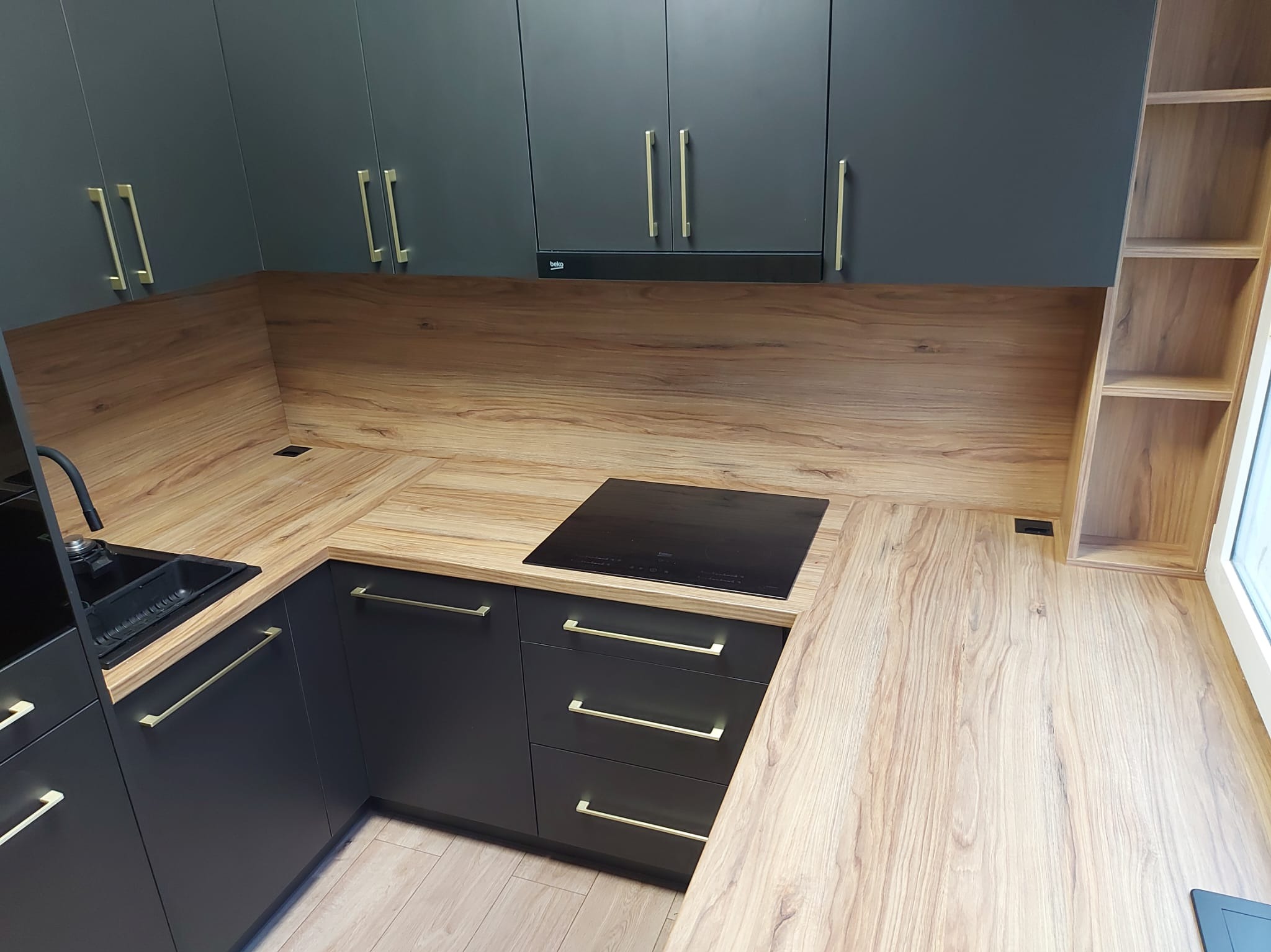 Nowoczesna kuchnia z ciemnymi szafkami i drewnianym blatem. Złote uchwyty dodają elegancji. Widoczna płyta indukcyjna i zlewozmywak.