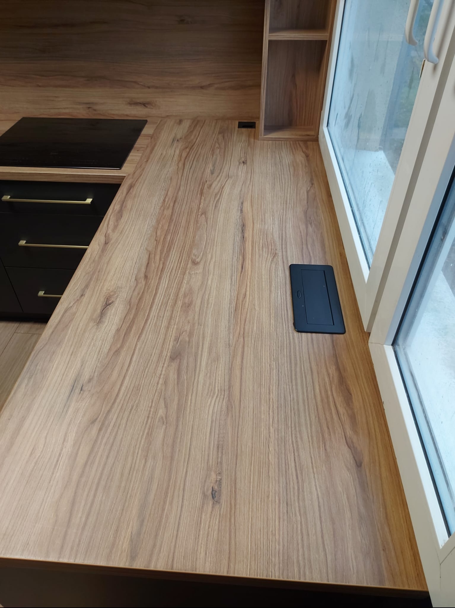 Drewniany blat kuchenny z czarną płytą indukcyjną i gniazdem wpuszczanym, widok z góry, oświetlenie naturalne z okna. Minimalistyczny design.