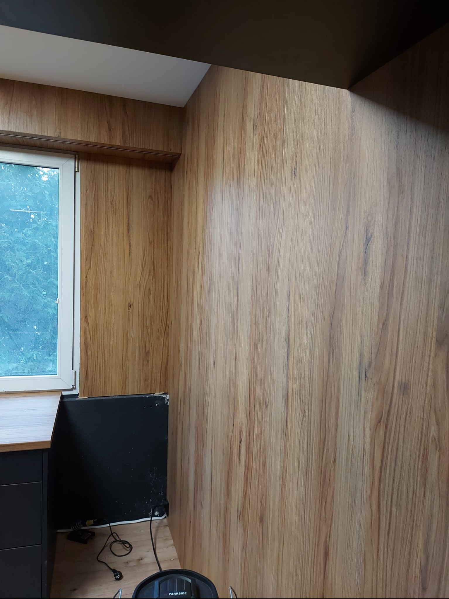 Wykończona ściana w okleinie drewnopodobnej, z widocznym fragmentem okna i blatu. Ujęcie z niskiego kąta, podkreślające fakturę i kolorystykę materiału.