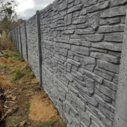 Długi, szary mur o fakturze kamienia z betonowych elementów, widoczny z bliskiej perspektywy, z ziemią i trawą u podstawy, pod pochmurnym niebem.
