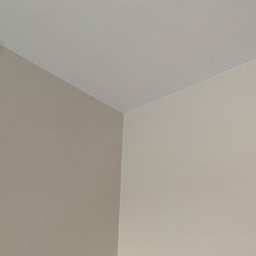 Majster Alex - Fragment rogu pokoju po malowaniu, gdzie ściany w odcieniach beżu spotykają się z białym sufitem, tworząc czyste i minimalistyczne połączenie.