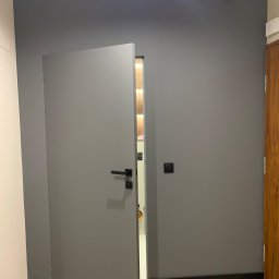 Majster Alex - Szara ściana po malowaniu z otwartymi szarymi drzwiami z czarną klamką i widocznym fragmentem szafy wnękowej wewnątrz.