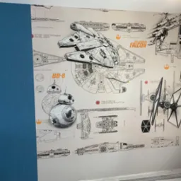 Ściana z tapetą przedstawiającą schematy statków kosmicznych z Gwiezdnych Wojen, w tym Millennium Falcon, BB-8, X-Wing i inne, częściowo zasłonięta przez pomalowaną na niebiesko ścianę z widocznym...