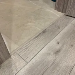 Połączenie ułożonych paneli podłogowych w odcieniach szarości z jasnymi płytkami ceramicznymi, widoczna krawędź szafki w kolorze paneli.