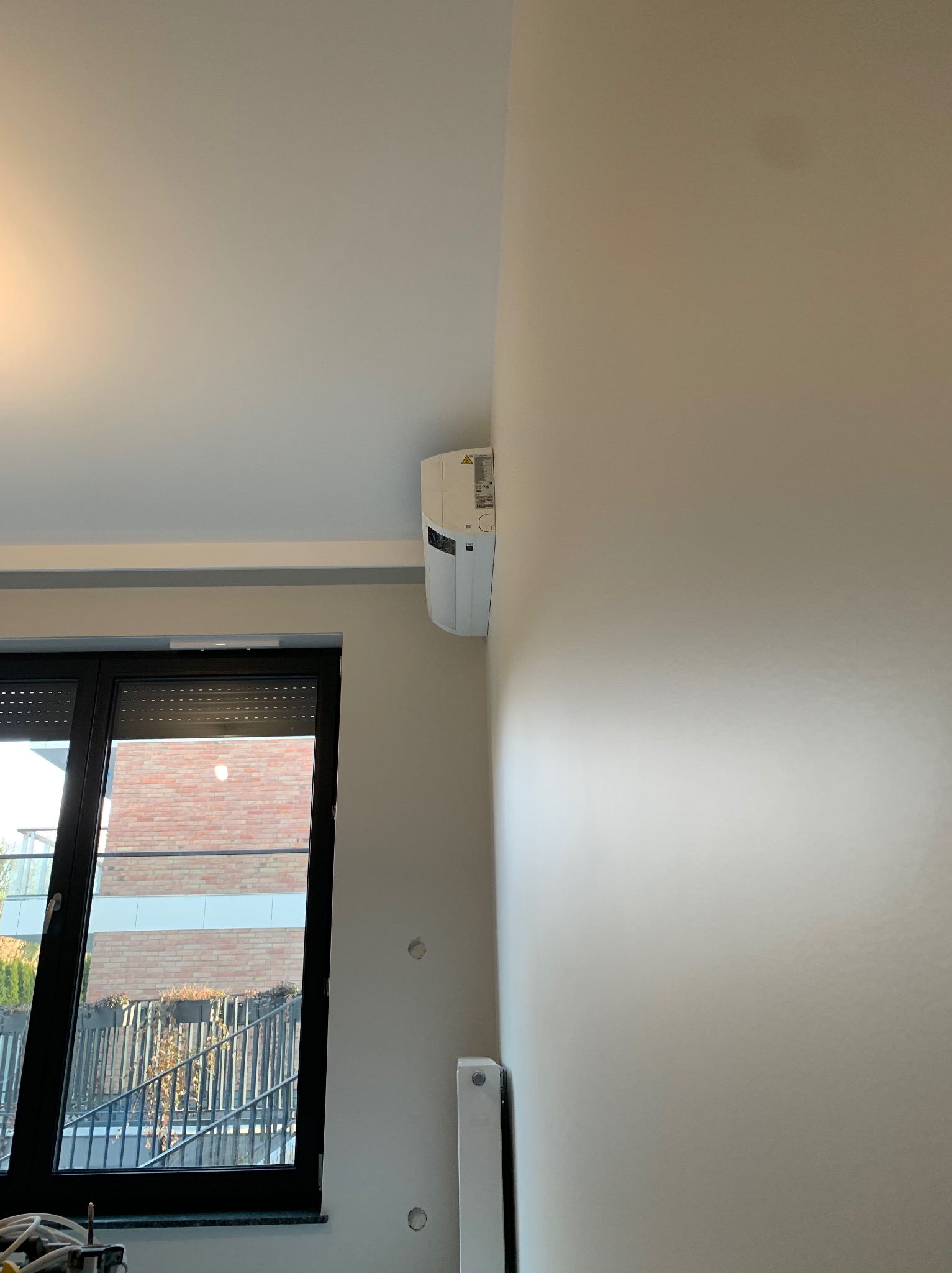 Wnętrze z zamontowanym klimatyzatorem na ścianie, widoczne otwory w ścianie przed malowaniem, okno z roletami zewnętrznymi.