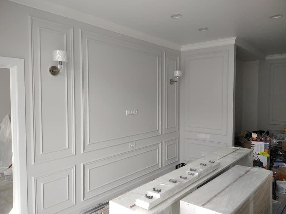 Eleganckie panele ścienne w kolorze złamanej bieli z dekoracyjnymi listwami i kinkietami, widoczne gniazdka elektryczne, prace w toku z zabezpieczonymi elementami wyposażenia wnętrz.