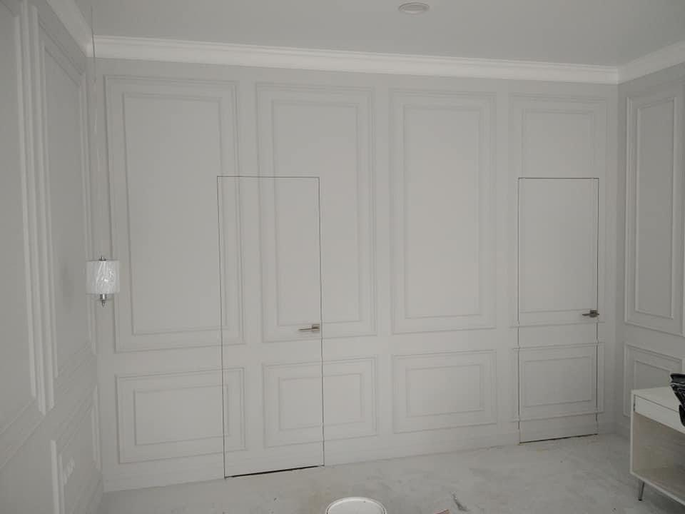 Eleganckie, szare wnętrze po remoncie z ozdobnymi panelami ściennymi, ukrytymi drzwiami z minimalistycznymi klamkami i listwami przypodłogowymi, widoczny fragment lampy ściennej i mebla.